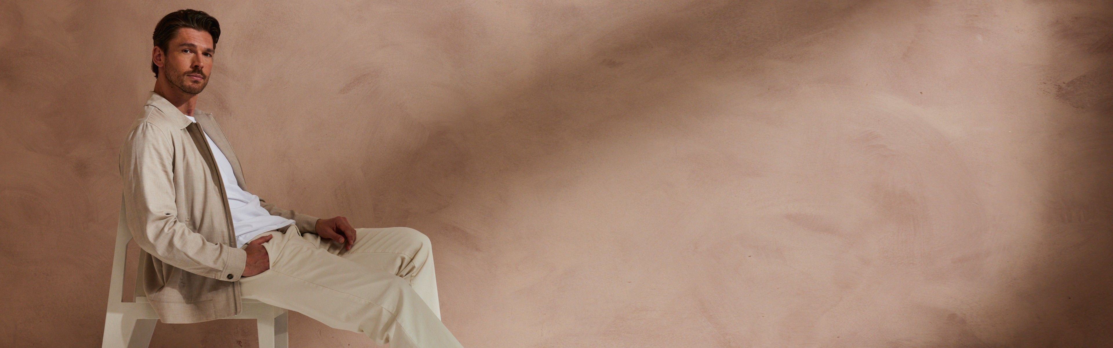 Man in beige jacket and pants sits on a chair against a brown textured background, looking at the camera.