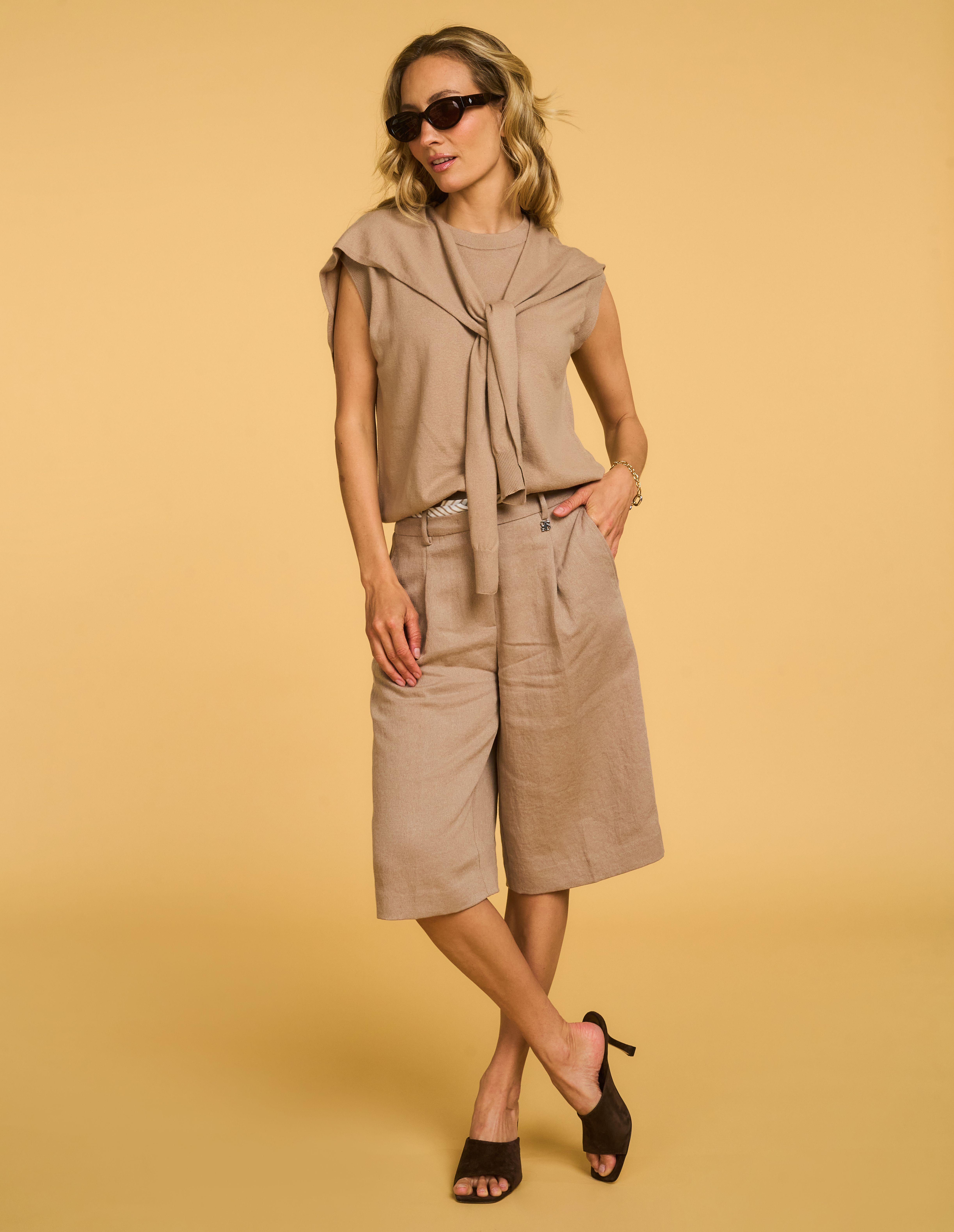 Person in a taupe outfit with a tied sweater, sunglasses, and heeled sandals poses against a beige background.