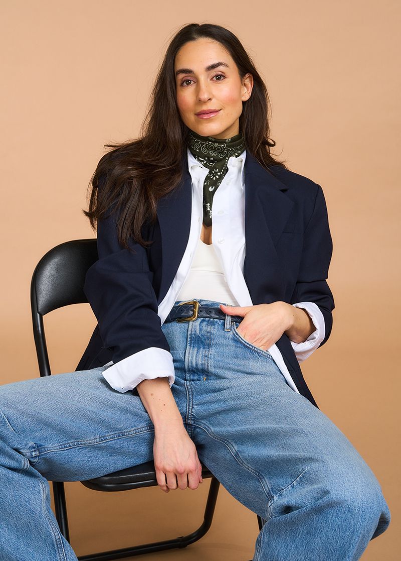 Person seated confidently, wearing a dark blazer, white shirt, green scarf, and blue jeans against a plain, neutral background.