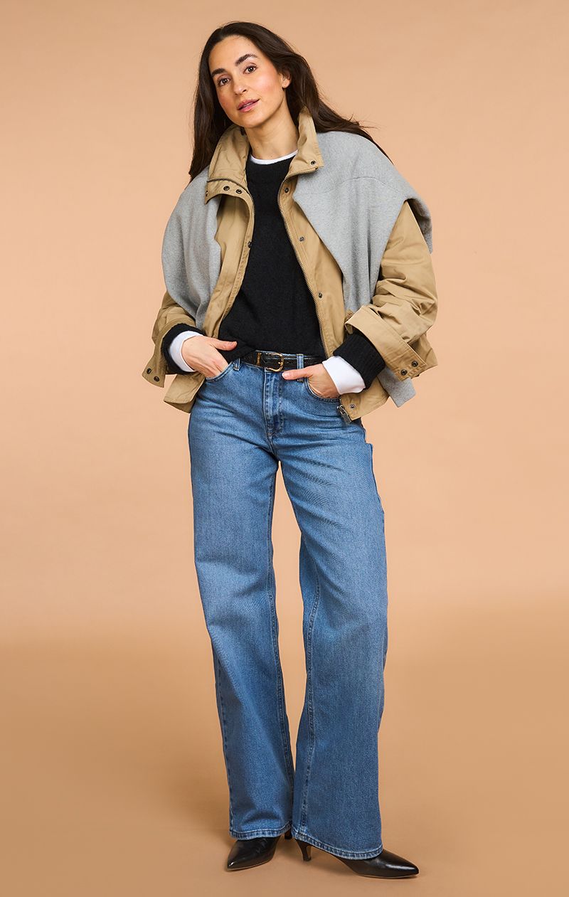 Woman wearing a layered outfit: beige jacket, gray scarf, black sweater, blue jeans, and black shoes, standing against a beige background.