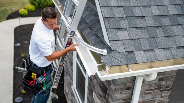 Person on a ladder installing a rain gutter on a house with gray roof shingles