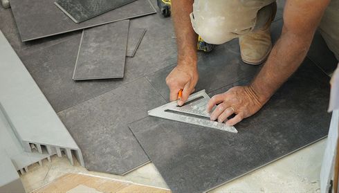 Person using a triangular ruler and pencil to measure gray tiles on a floor, preparing for installation