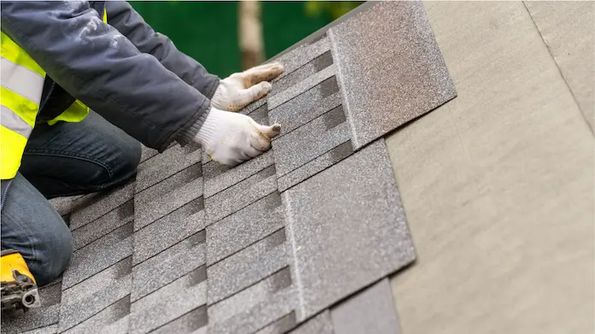Person in gloves and protective gear installing asphalt shingles on a roof.