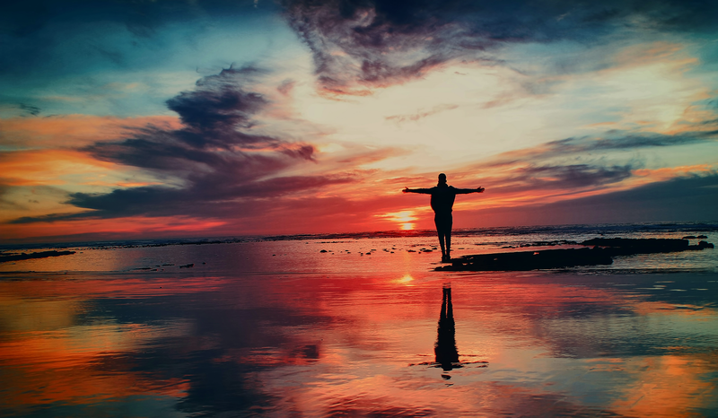 Silhouette of a person with outstretched arms standing on a reflective beach at sunset, with vibrant red and blue sky.
