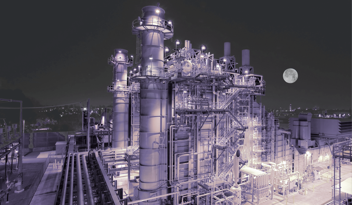 Industrial plant at night with illuminated towers and structures, under a clear sky with a visible full moon in the background.