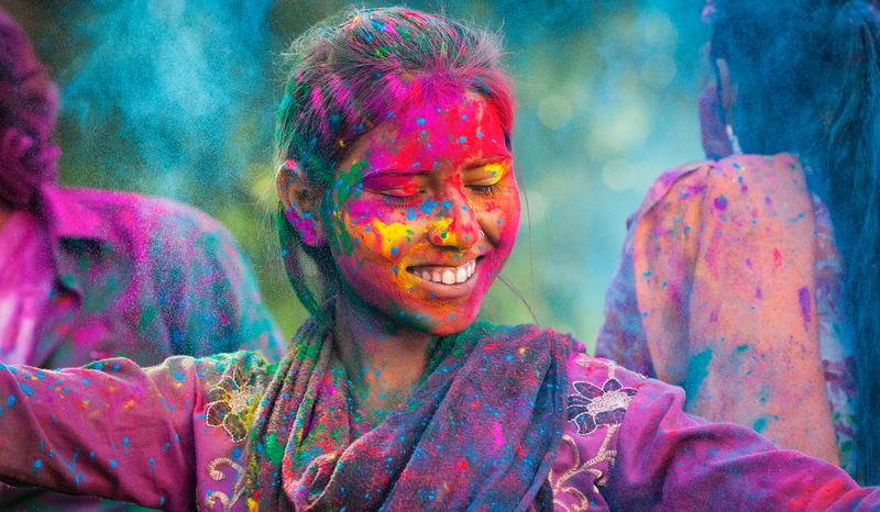 A smiling person covered in vibrant colored powders during a Holi celebration, with eyes closed and colorful background.