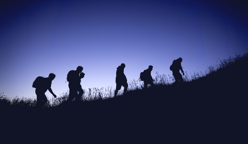 Silhouetted hikers with backpacks walking uphill against a twilight sky, surrounded by grass.