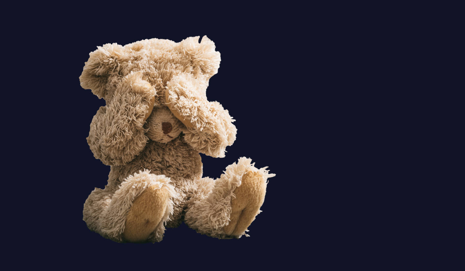 Fluffy teddy bear sitting with its paws covering its eyes against a dark background.