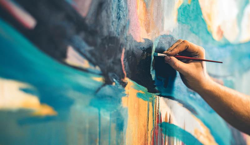 Close-up of a hand painting a colorful abstract mural with a brush, featuring vibrant blues, oranges, and pinks.