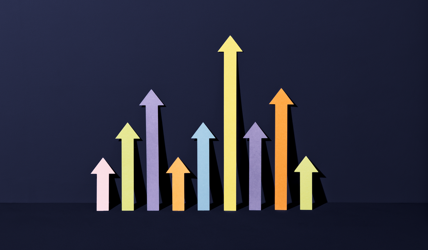 Colorful paper arrows of varying heights pointing upward against a dark background, symbolizing growth or progress.