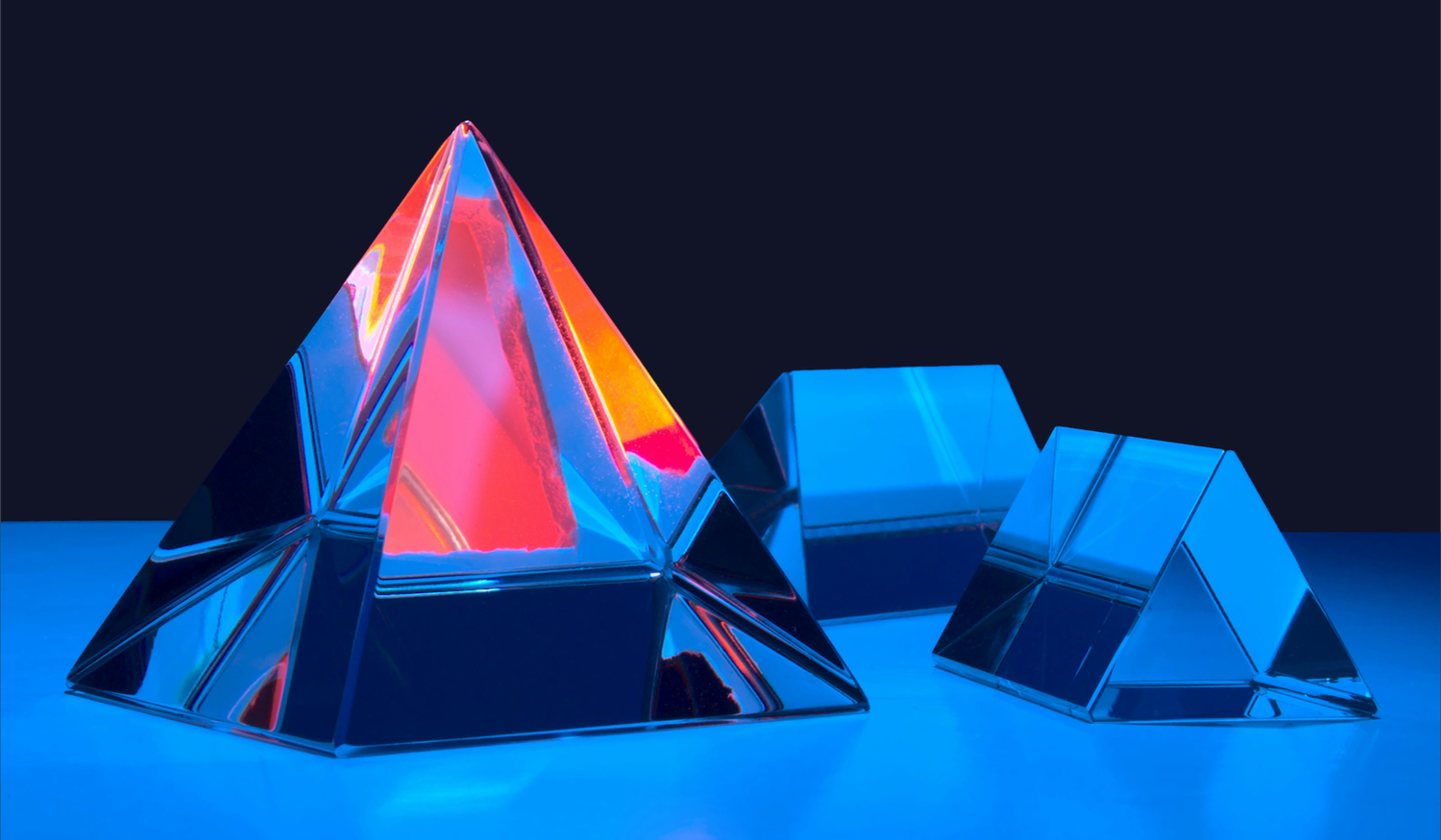 Three glass pyramids with reflective surfaces, one glowing with red and orange hues, against a dark blue background.