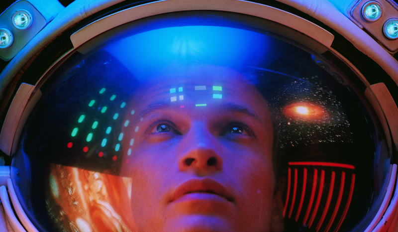 Person inside a futuristic helmet with colorful lights reflecting on the visor, gazing upwards.