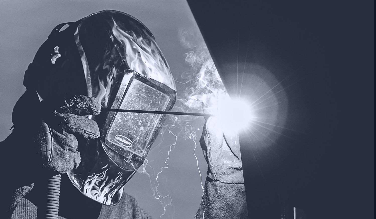 Welder in protective gear works with bright sparks and smoke, wearing a helmet with flame design, against a dark background.