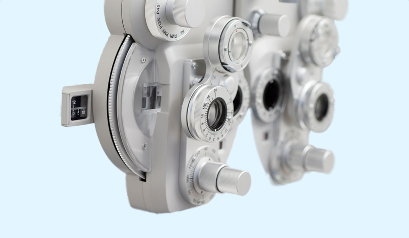 Close-up of a phoropter, an optical device with lenses and dials, used for eye exams against a light blue background.