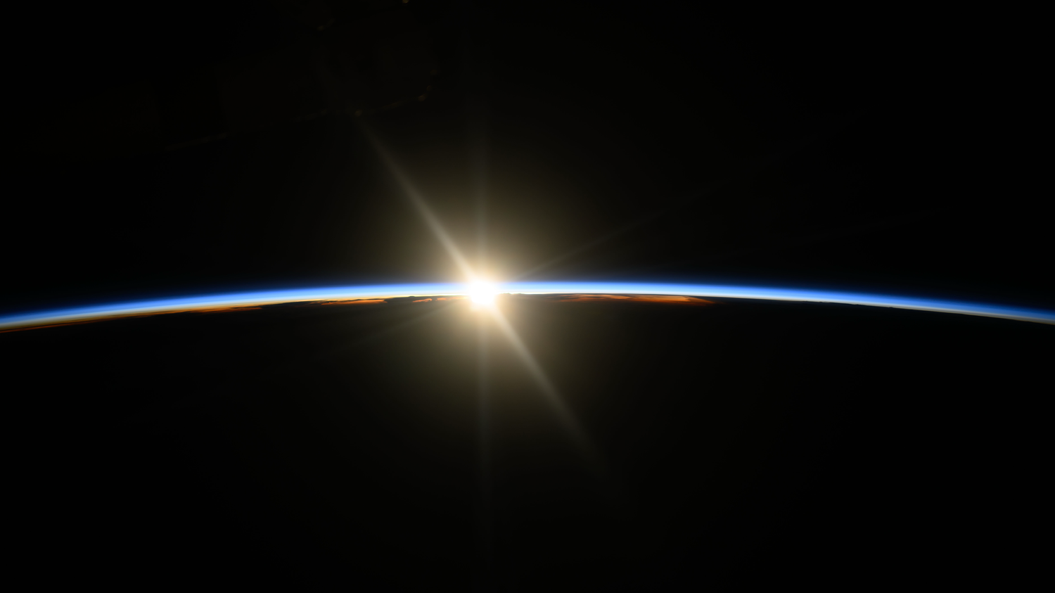 Sunrise seen from space, with the sun peeking over Earth's horizon, casting a bright glow and illuminating the thin blue atmosphere.
