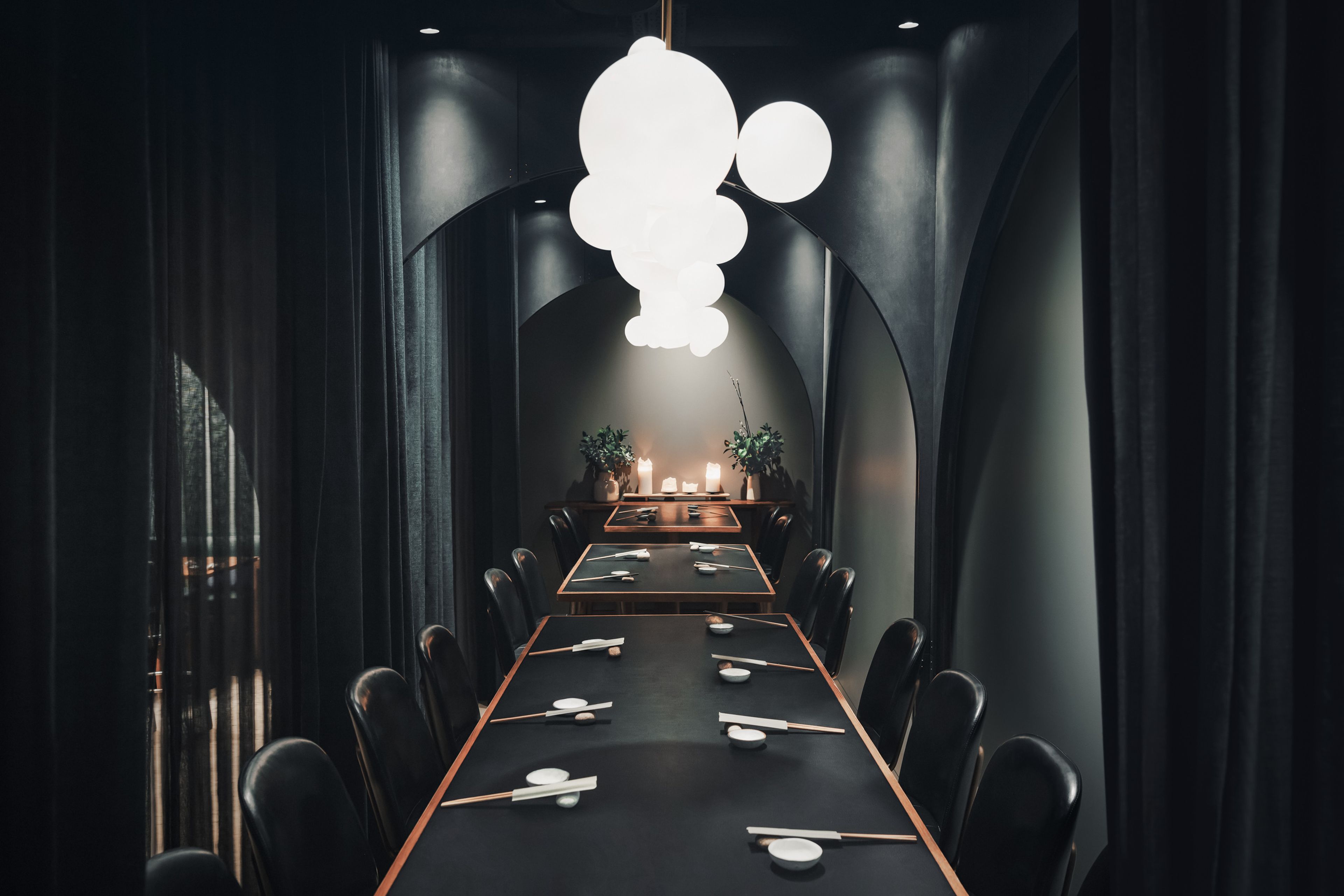 Elegant dining room with long tables set for a meal, surrounded by dark curtains and illuminated by spherical pendant lights.