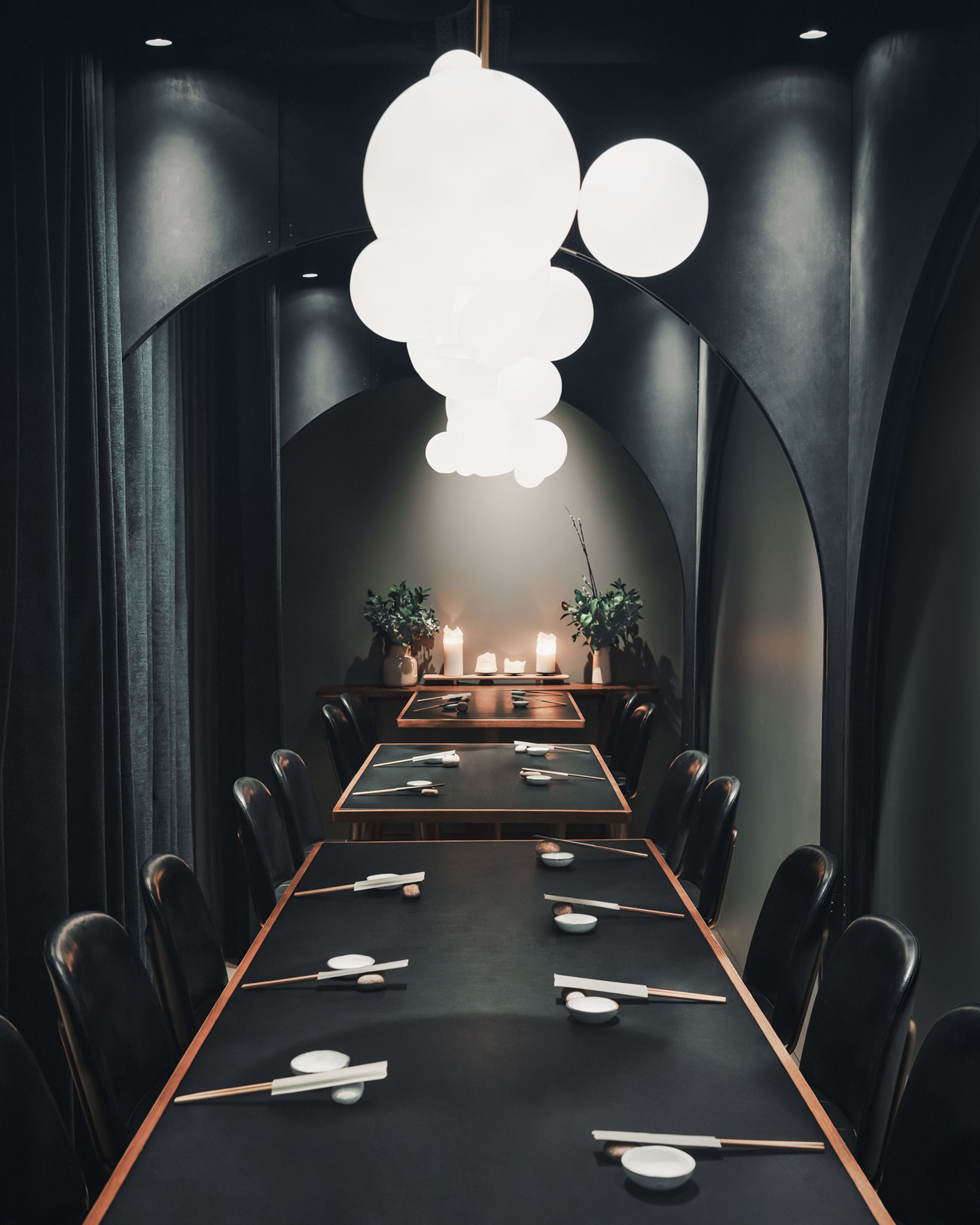 Elegant dining room with long tables set for a meal, surrounded by dark curtains and illuminated by spherical pendant lights.