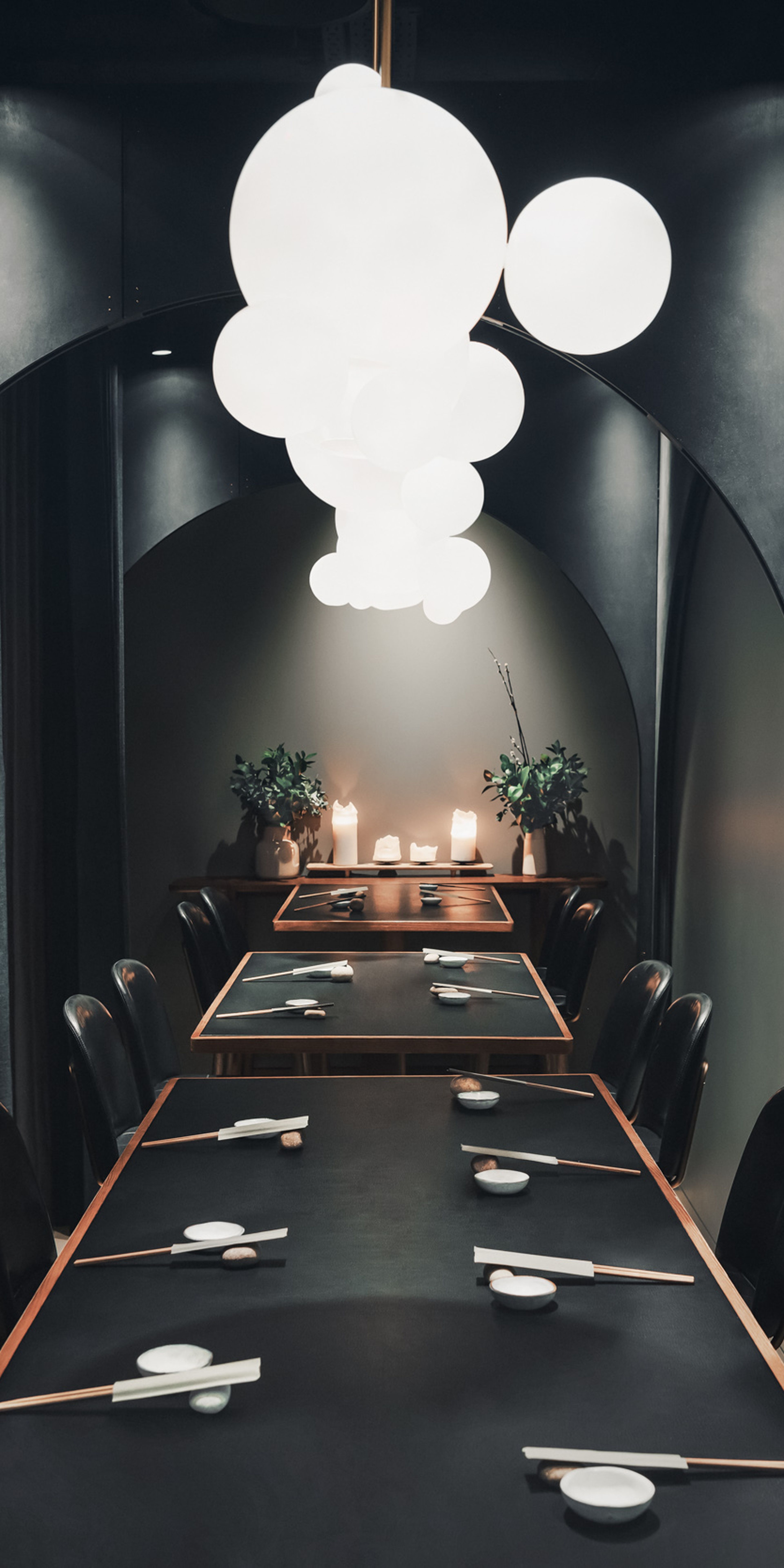 Elegant dining room with long tables set for a meal, surrounded by dark curtains and illuminated by spherical pendant lights.