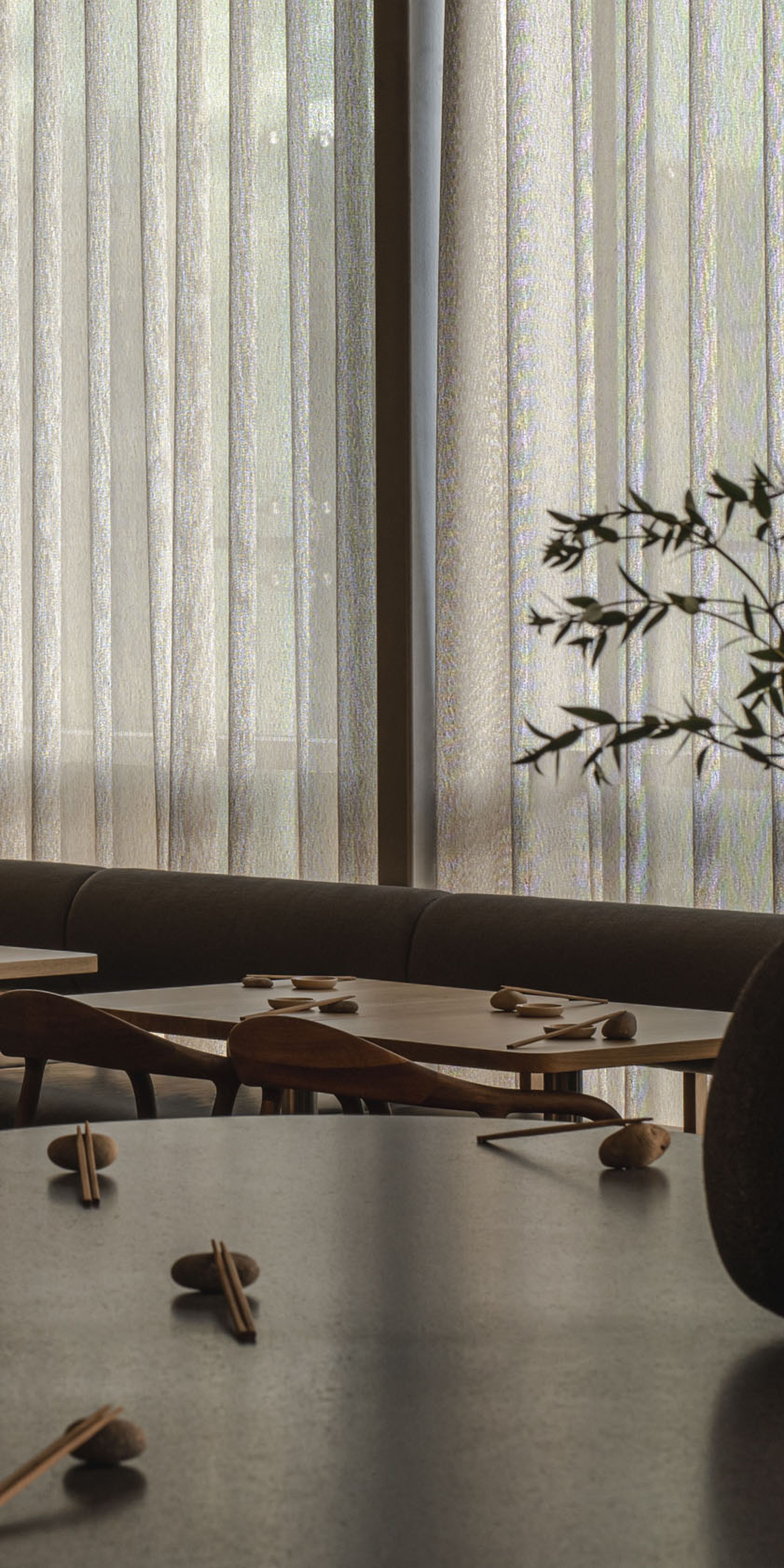Modern dining area with wooden chairs, tables set with utensils, and vases with branches on a counter. Soft light filters through sheer curtains.