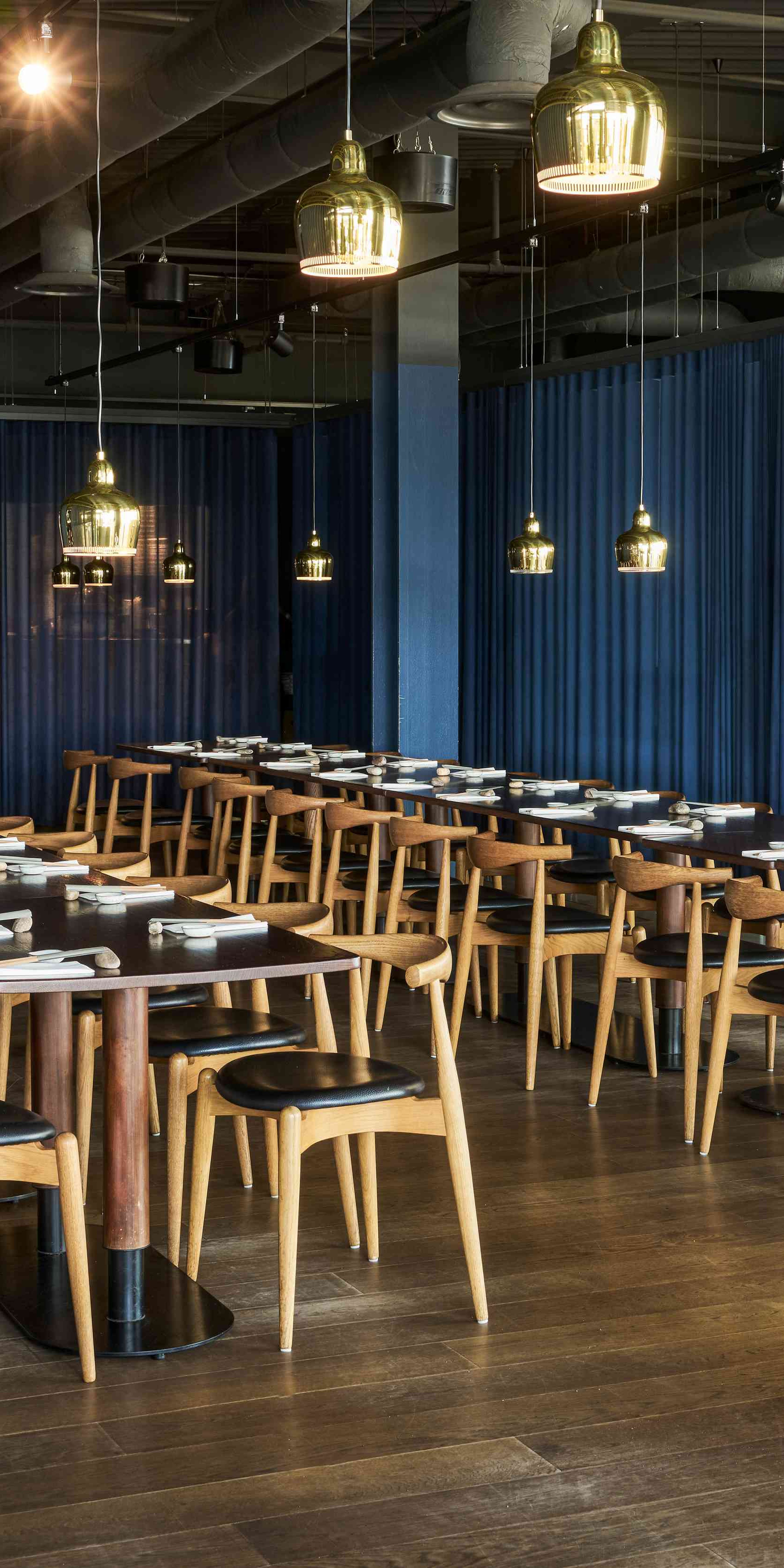 Modern restaurant interior with wooden tables and chairs, blue curtains, and hanging pendant lights, creating an elegant dining atmosphere.
