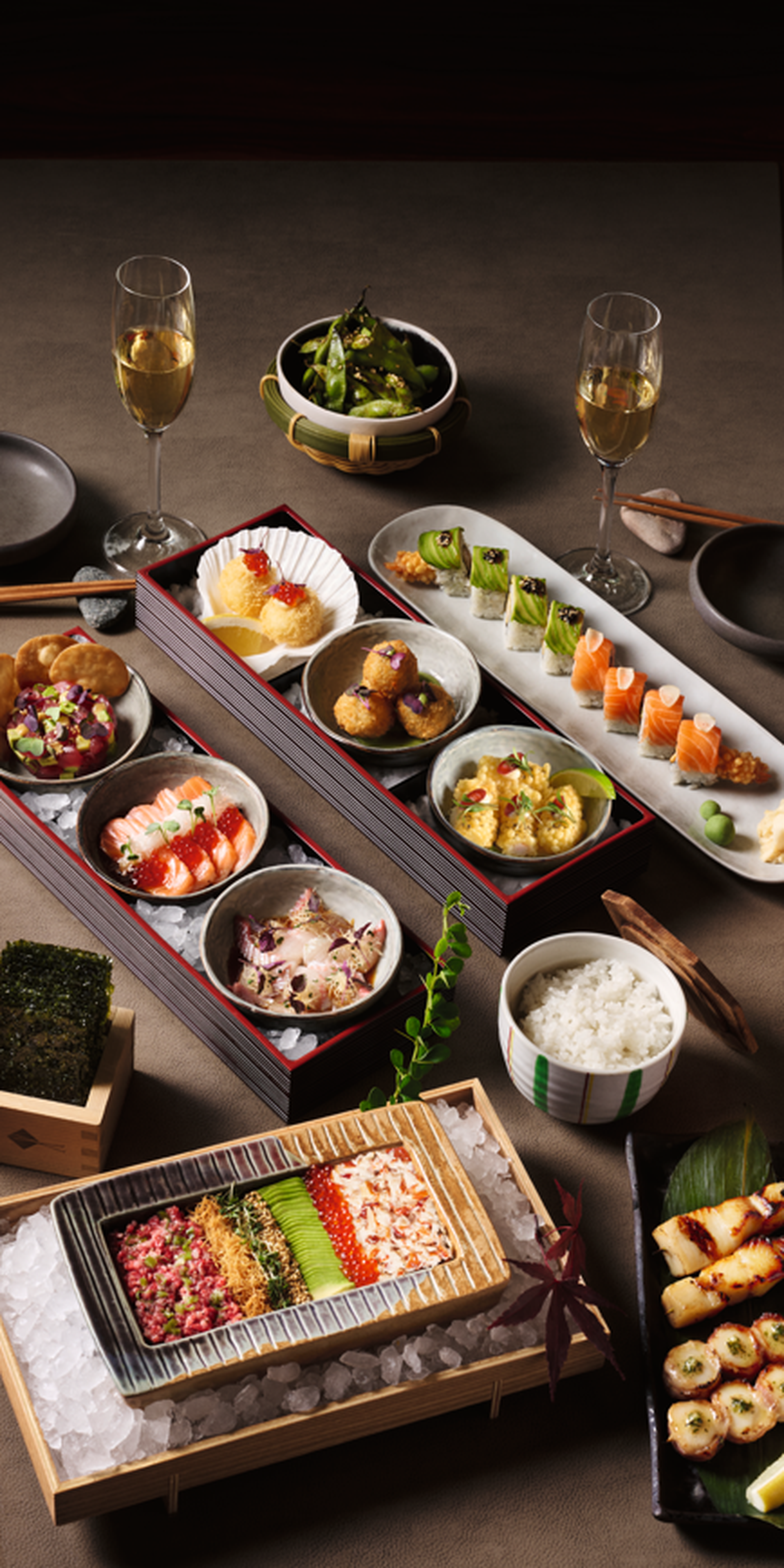 A variety of elegantly arranged Japanese dishes, including sushi, skewers, and salads, on a wooden table with two glasses of champagne.