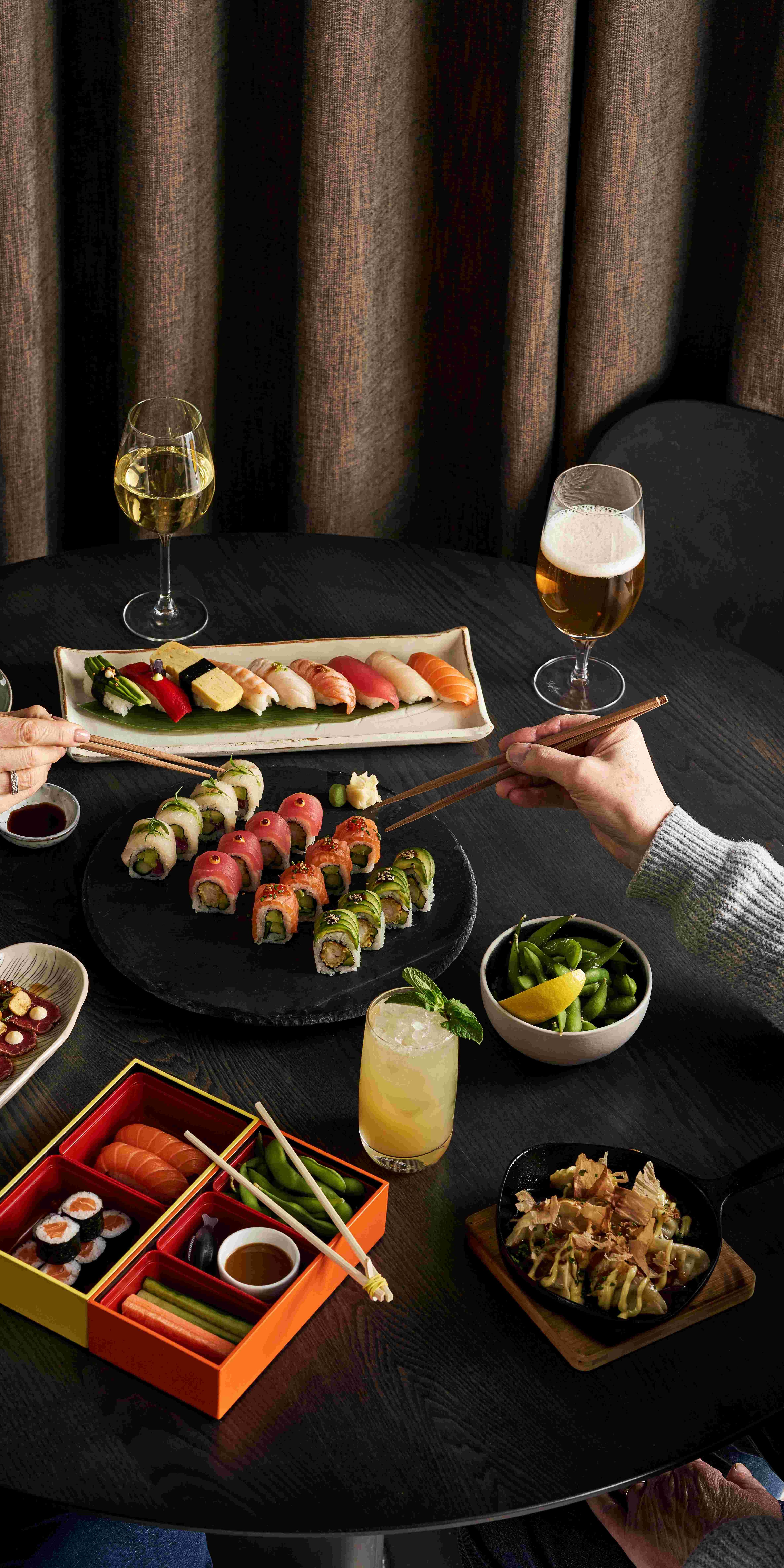 A table with assorted sushi, sashimi, edamame, and drinks. Two people use chopsticks to pick sushi from shared plates.