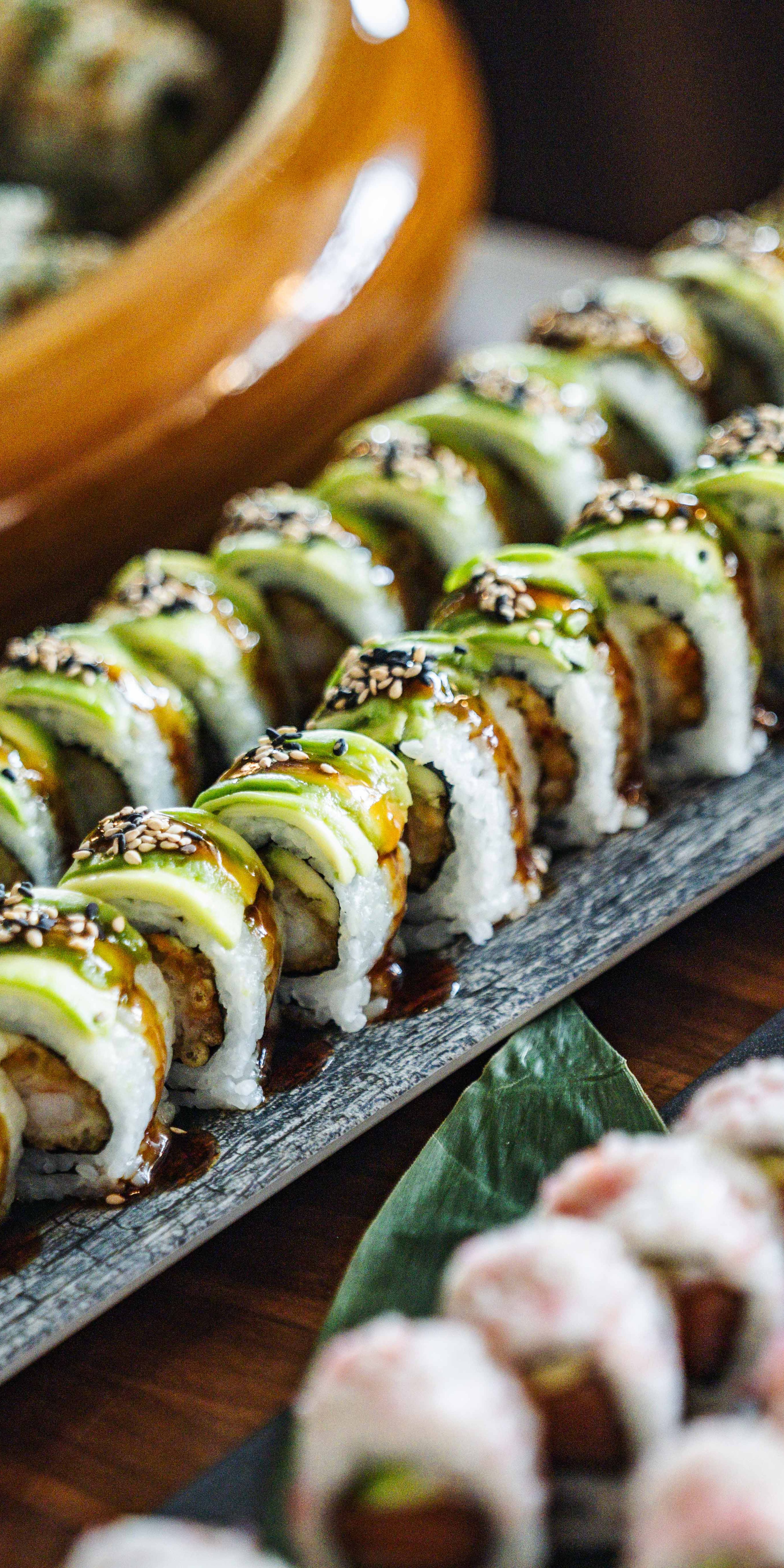 Assorted sushi rolls on a platter, featuring avocado-topped rolls with sesame seeds and pink-tinted rolls with visible fillings.