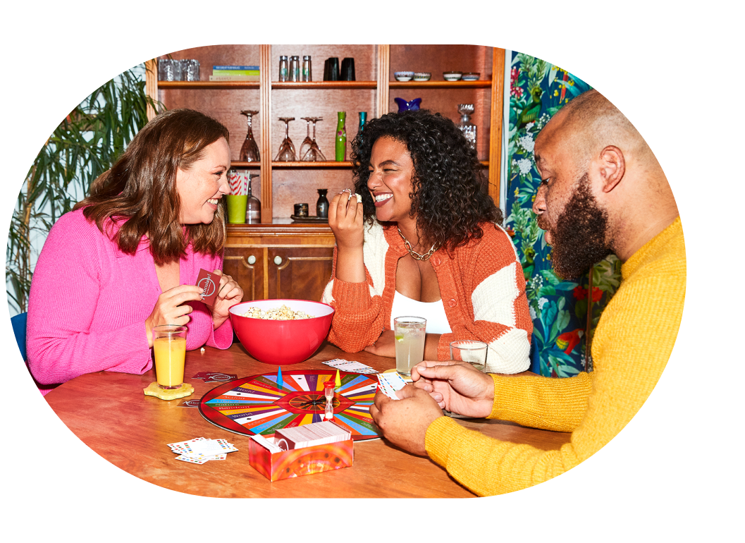 Three friends laughing and playing a board game with popcorn and drinks.