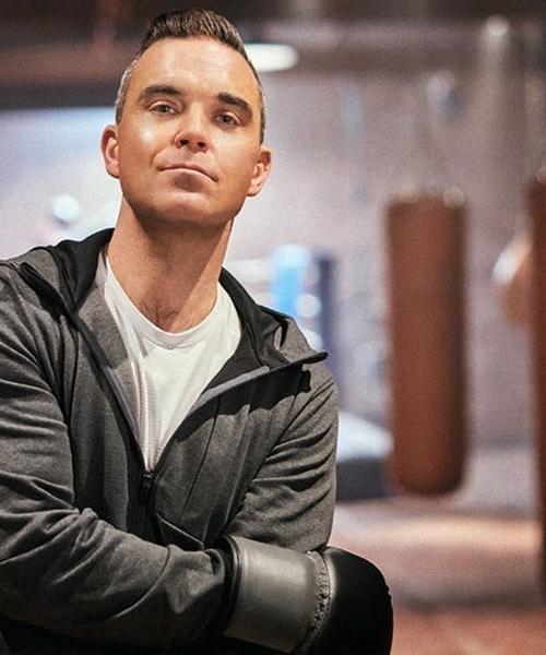 Robbie Williams in boxing gloves, looking determined in a gym setting.