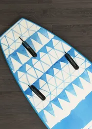 A surfboard with a blue and white geometric pattern and three black fins, resting on a dark wood floor.