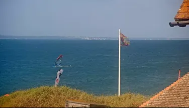 Windsurfer on a blue ocean, with colorful sails, viewed from a grassy hill with flags and tiled rooftop in the foreground.
