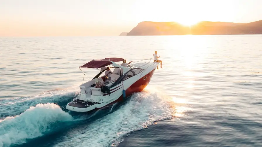 A boat with a maroon canopy cruises on calm water at sunset, with one person sitting at the bow and mountains in the background.