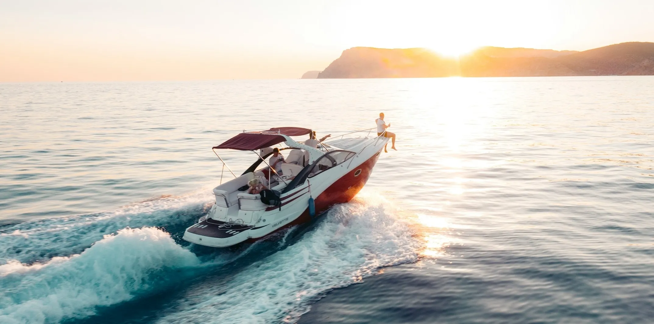 A boat with a maroon canopy cruises on calm water at sunset, with one person sitting at the bow and mountains in the background.
