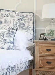 Elegant bedroom with a floral-patterned headboard and pillow, white bedding, wooden nightstand with lamp, framed photo, and leafy decor.