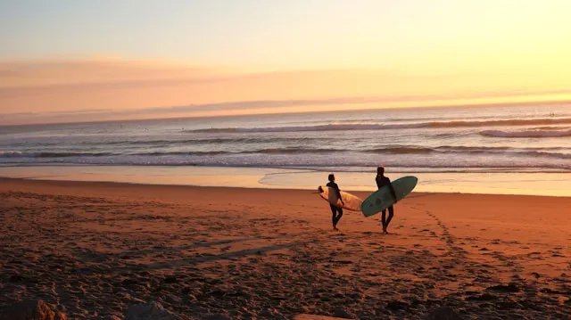Two surfers walk along a sandy beach with surfboards at sunset, the sky glowing in warm hues over the ocean waves.