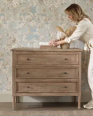 Woman arranging items on a wooden dresser against a floral wallpaper, dressed in a light outfit and sneakers.