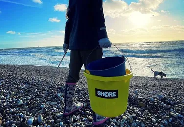 Person in boots holds a yellow bucket and picker on a rocky beach, with the ocean and a dog running in the background under a bright sky.