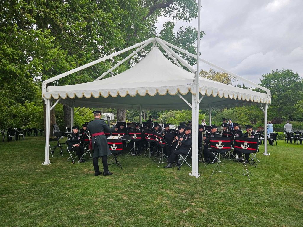 A band performs under a large white tent in a grassy park, with a conductor in uniform leading them. Trees and sky are visible in the background.