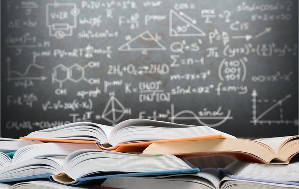 Intensivkurs oder Ferienkurs Nachhilfe Mathematik Offene Bücher, die vor einer Tafel mit mathematischen Gleichungen und geometrischen Diagrammen gestapelt sind.