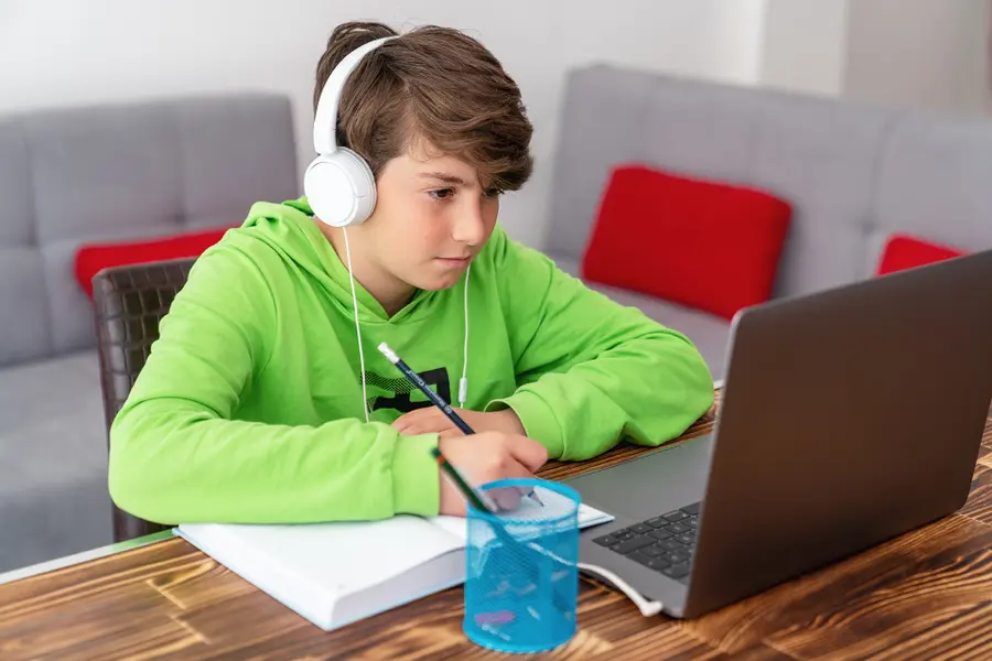 Ein Junge in einem grünen Kapuzenpulli mit Kopfhörern sitzt an einem Tisch und ist auf seinen Laptop konzentriert. Ein Notizbuch und ein Bleistift liegen daneben.