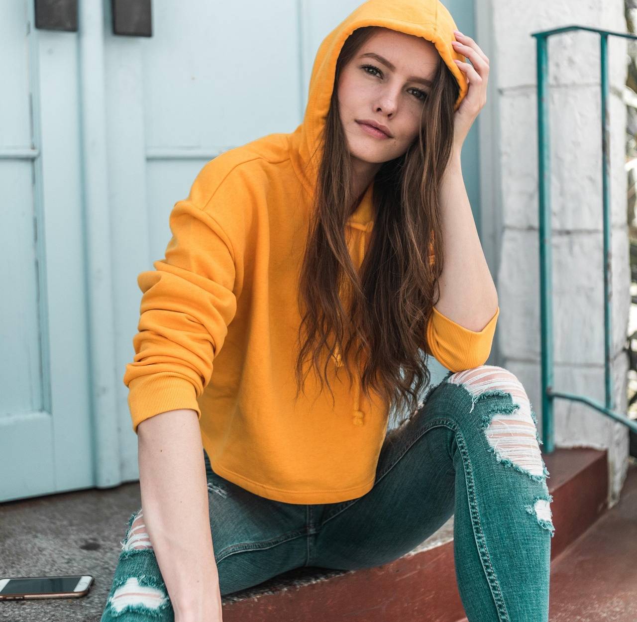 A person in a yellow hoodie and ripped jeans sitting on steps, with a phone nearby, in front of a light blue door.
