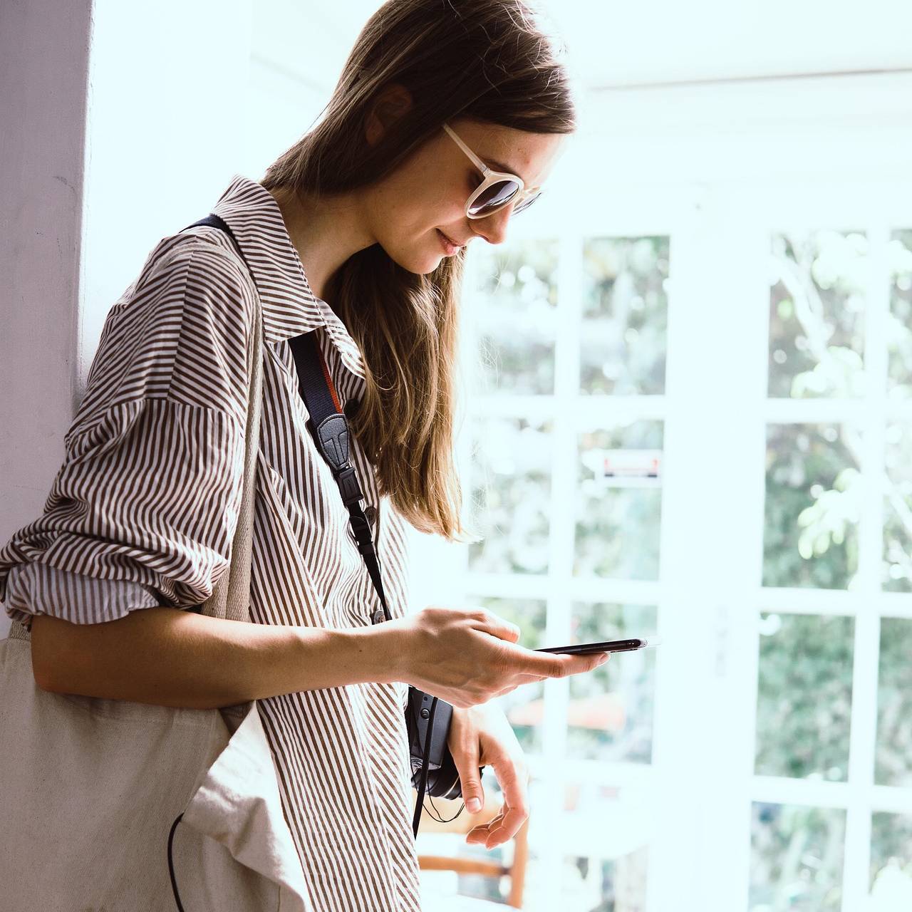 Woman in a striped shirt and sunglasses, holding a phone and a fabric bag, standing by a window with natural light streaming in.