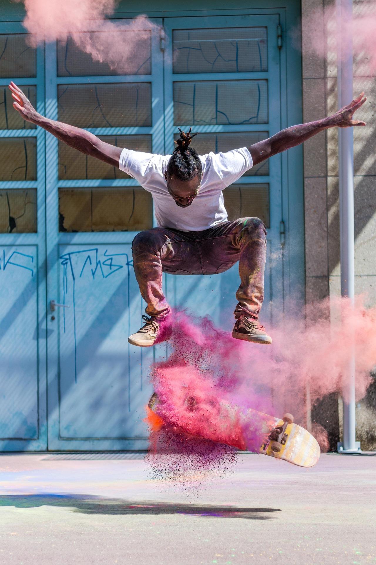 Skateboardåkare i luften med uppochnedvänd bräda medan rosa och lila pulver sprängs runt scenen framför blå dörrar.
