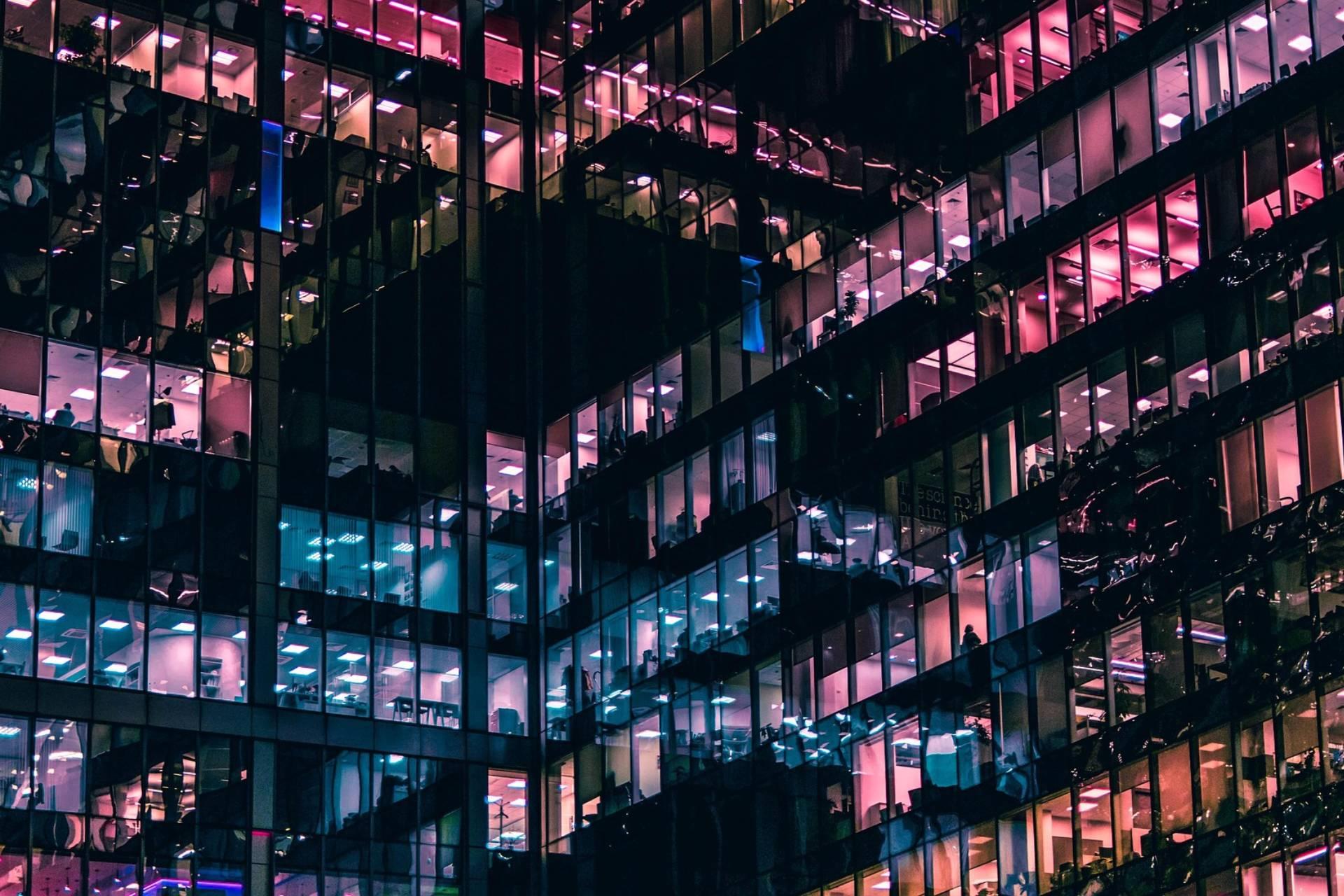 Nattvy över en modern glasbyggnad med upplysta kontor i rosa och blå nyanser, som skapar ett livligt, geometriskt mönster.