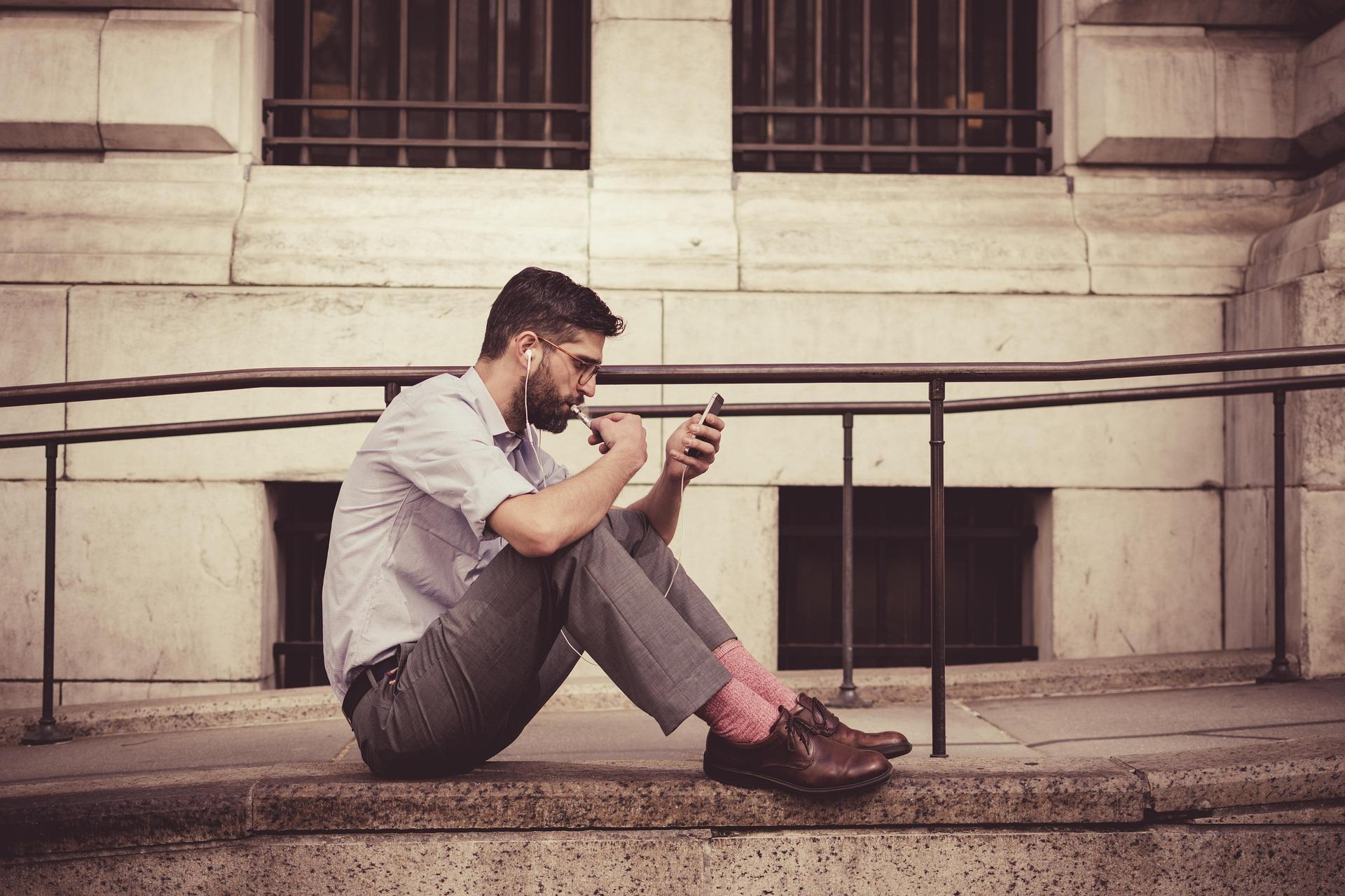 A man sitting on a ledge, wearing headphones, looking at his phone, dressed in a light shirt, gray pants, pink socks, and brown shoes.