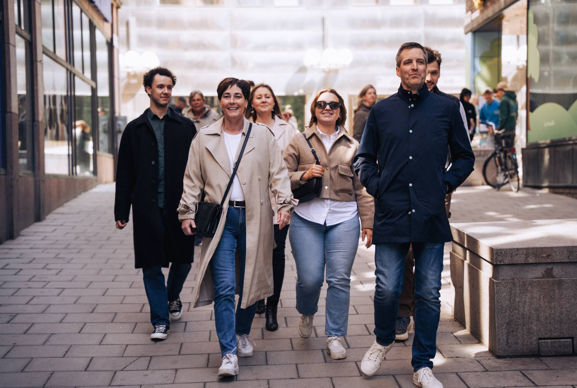 En grupp människor går självsäkert nerför en stadsgata, iklädda avslappnade jackor och jeans, med moderna byggnader i bakgrunden.