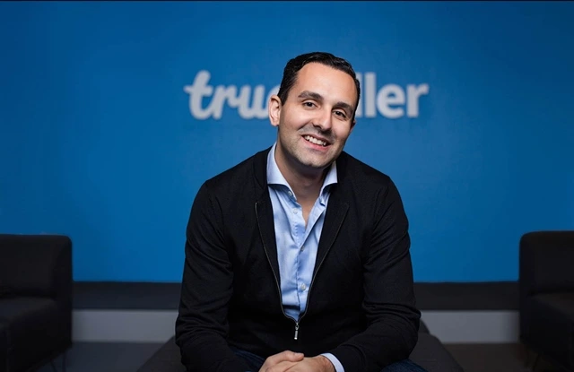 Alan Mamedi in business attire smiling while sitting in front of a Truecaller logo