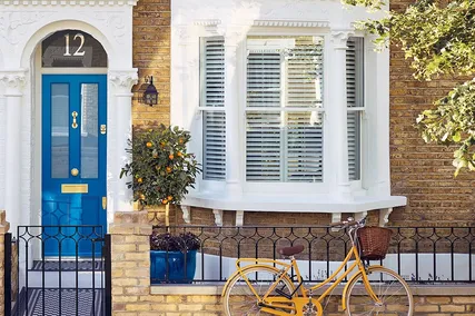 House with bespoke blue door, brass number 12 and Banham premium locks, white-trimmed bay window, yellow bike with a basket, and potted plant by a brick fence.