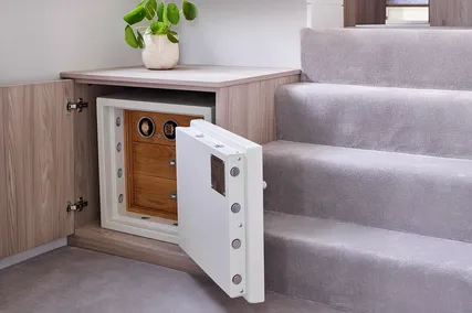 A safe is built into a carpeted staircase, with an open door revealing its interior. A small plant sits on a nearby wooden surface.
