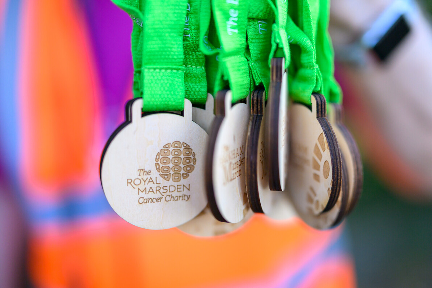 Close-up of wooden medals with green ribbons, featuring "The Royal Marsden Cancer Charity" logo, held by a person in an orange vest.